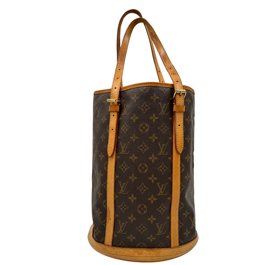 Front di un bucket Louis Vuitton in canvas marrone monogram con finiture in vacchetta naturale, manici regolabili con fibbie e parti metalliche dorate.