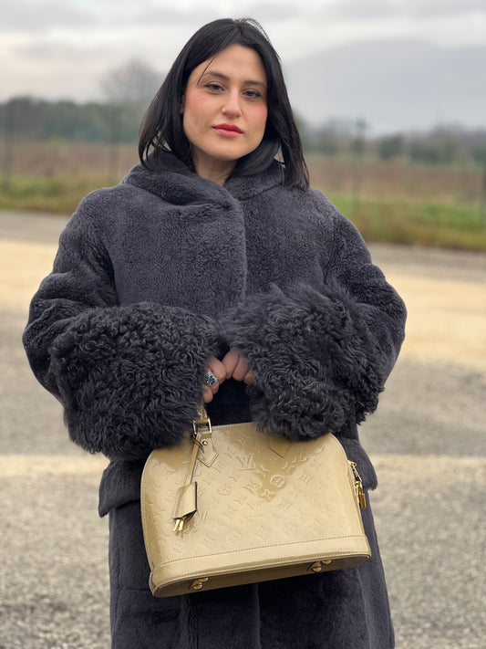 Ragazza che indossa una borsa Alma PM Louis Vuitton in pelle Vernis color noisette con parti metalliche dorate e doppi manici stondati; da portare a mano. Completa di lucchetto e chiavi.