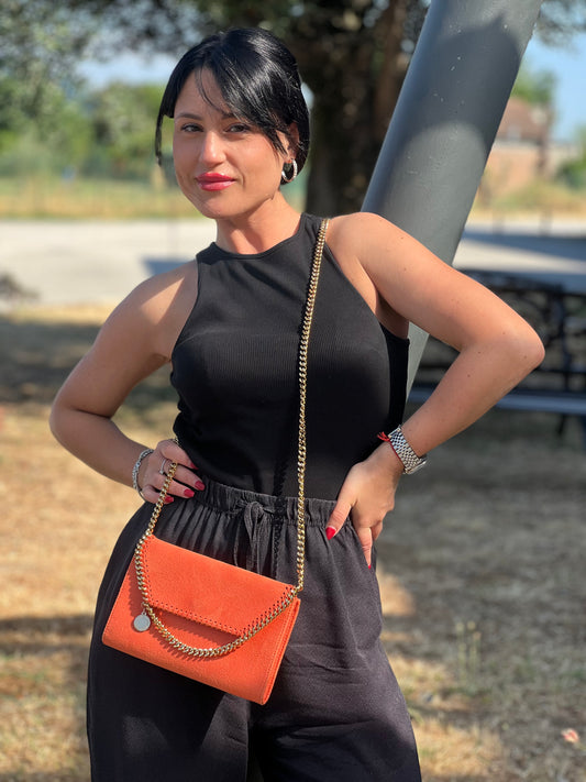 Modella all’aperto con completo nero smanicato e pantaloni larghi, che indossa a spalla una borsa a tracolla arancione in suede con catena dorata decorativa e medaglietta metallica.