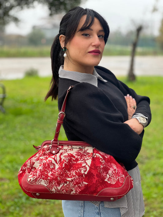 Ragazza che indossa Borsa Miu Miu in tessuto jacquard rosso e beige in trama floreale, con finiture in pelle rosse e parti metalliche argentate. Presenta doppi manici leggermente regolabili e chiusura con zip. Indossabile a spalla.