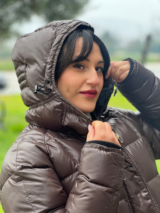 Ragazza che indossa un piumino Moncler in tessuto lucido color marrone, con parti metalliche effetto renio, in 100% poliammide, caratterizzato da imbottitura trapuntata, chiusura con zip e bottoni logati, cappuccio regolabile e polsini interni a coste per una maggiore protezione dal freddo.