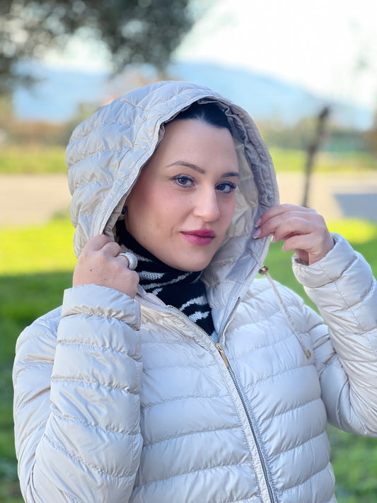 Ragazza che indossa un piumino Max Mara The Cube in 100% poliammide trapuntato, colore avorio perla. Modello con cappuccio, coulisse regolabile in vita e dettagli metallici dorati. Chiusura con zip.