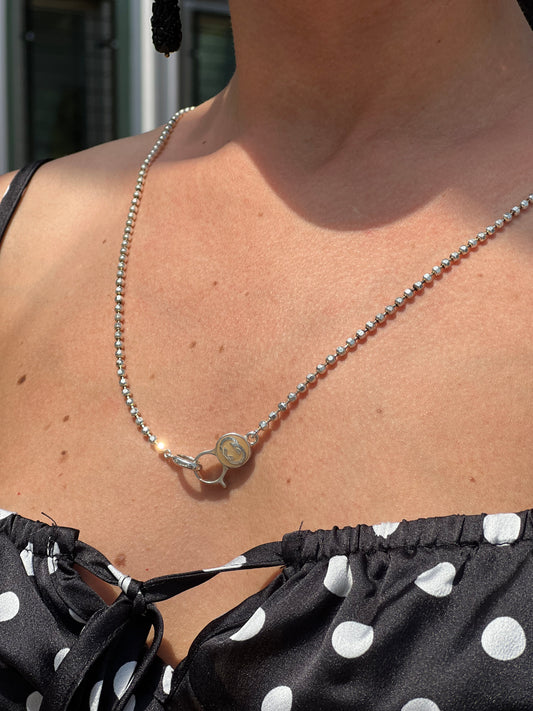 Ragazza che indossa una collana Gucci in argento 925 con catena a sfere sfaccettate pendente a moschettone rotondo che presenta il logo "GG" intrecciato smaltato su fondo marrone/beige. Munita di sacchetto anti polvere e cartellino.