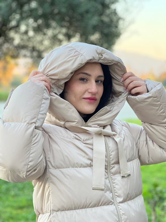 Ragazza che indossa piumino Herno in 100% poliestere color sabbia. Presenta una linea leggermente svasata, cappuccio ampio e avvolgente con coulisse decorativa, chiusura frontale con zip.