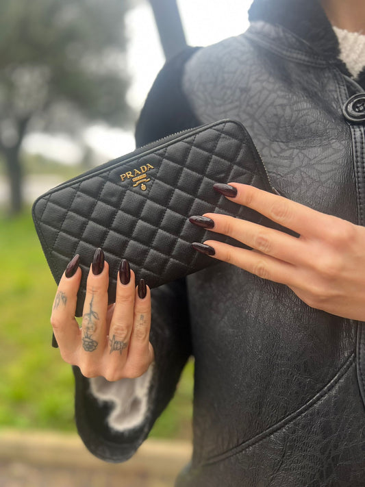 Ragazza che indossa un Portafoglio Prada in pelle trapuntata nera con parti metalliche dorate munito di vari scomparti, e rifinito con logo lettering sul front. Originale, usato, di lusso, in ottime condizioni.