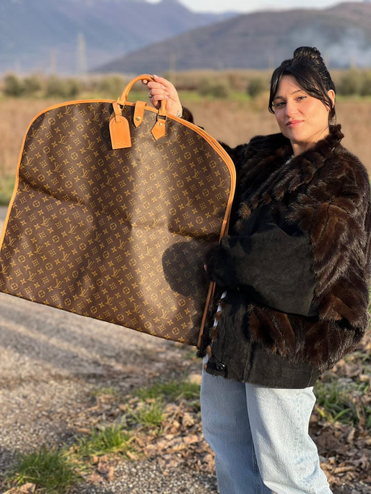 Ragazza che indossa un Porta abiti in canvas marrone monogram con finiture in vacchetta naturale e parti metalliche dorate; munita di due manici stondati. Completa di due lucchetto, chiavi, tag porta address e una dustbag. Originale, usata, di lusso, in ottime condizioni.