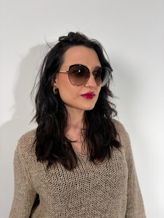 Ragazza che indossa un paio di occhiali da sole Prada caratterizzati da una montatura nera lucida con lenti sfumate grigie, asticelle maculate e hardware dorato, usati, originali, second hand, in ottime condizioni.