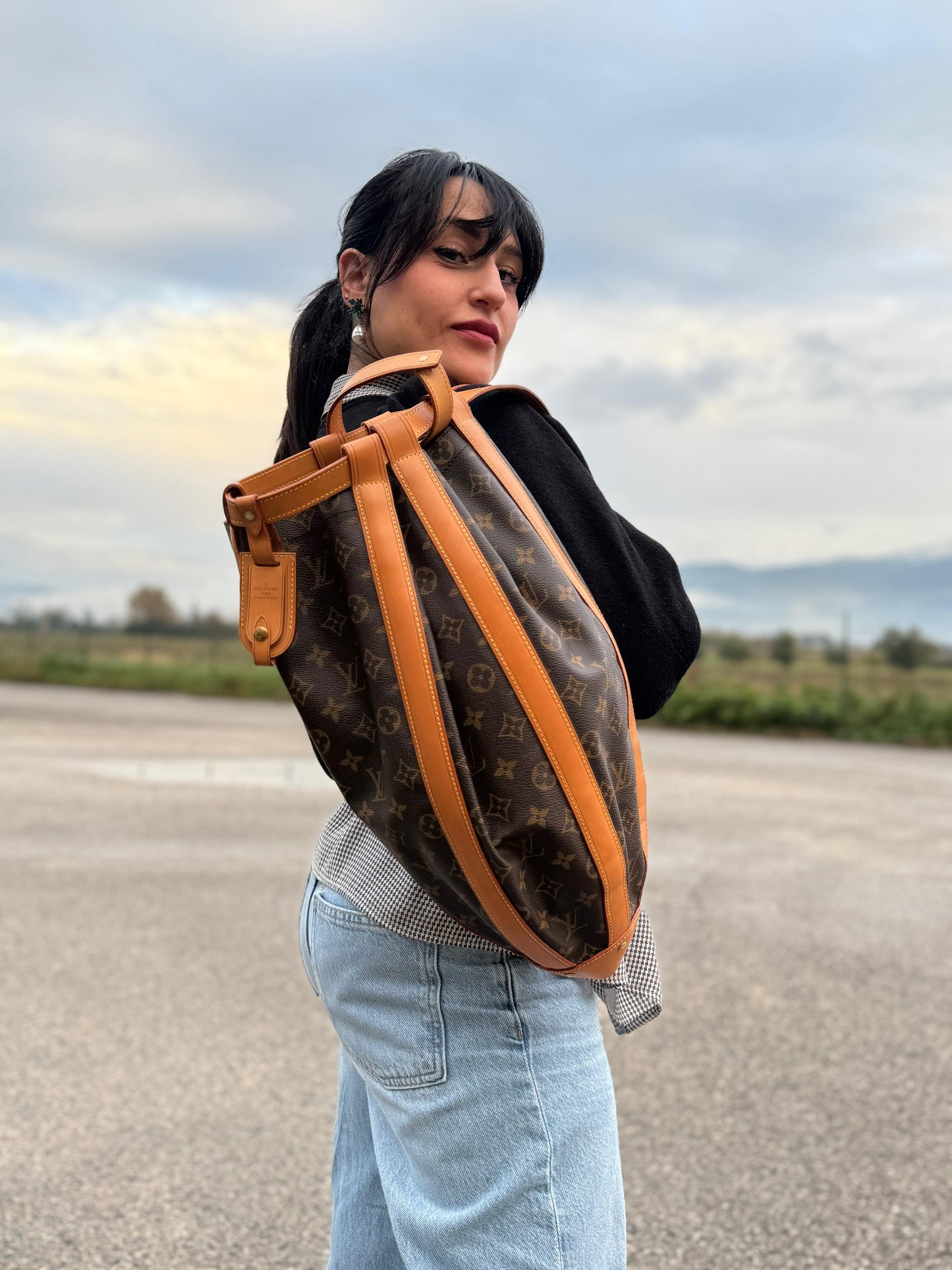Ragazza che mostra una borsa Louis Vuitton limited edition Romeo Gigli, in canvas Monogram e pelle naturale. Struttura cilindrica con rinforzi in pelle e finiture metalliche dorate. Chiusura superiore con coulisse. Spallaccio in pelle regolabile. Dotata di dustbag e manico amovibile. Indossabile a spalla.