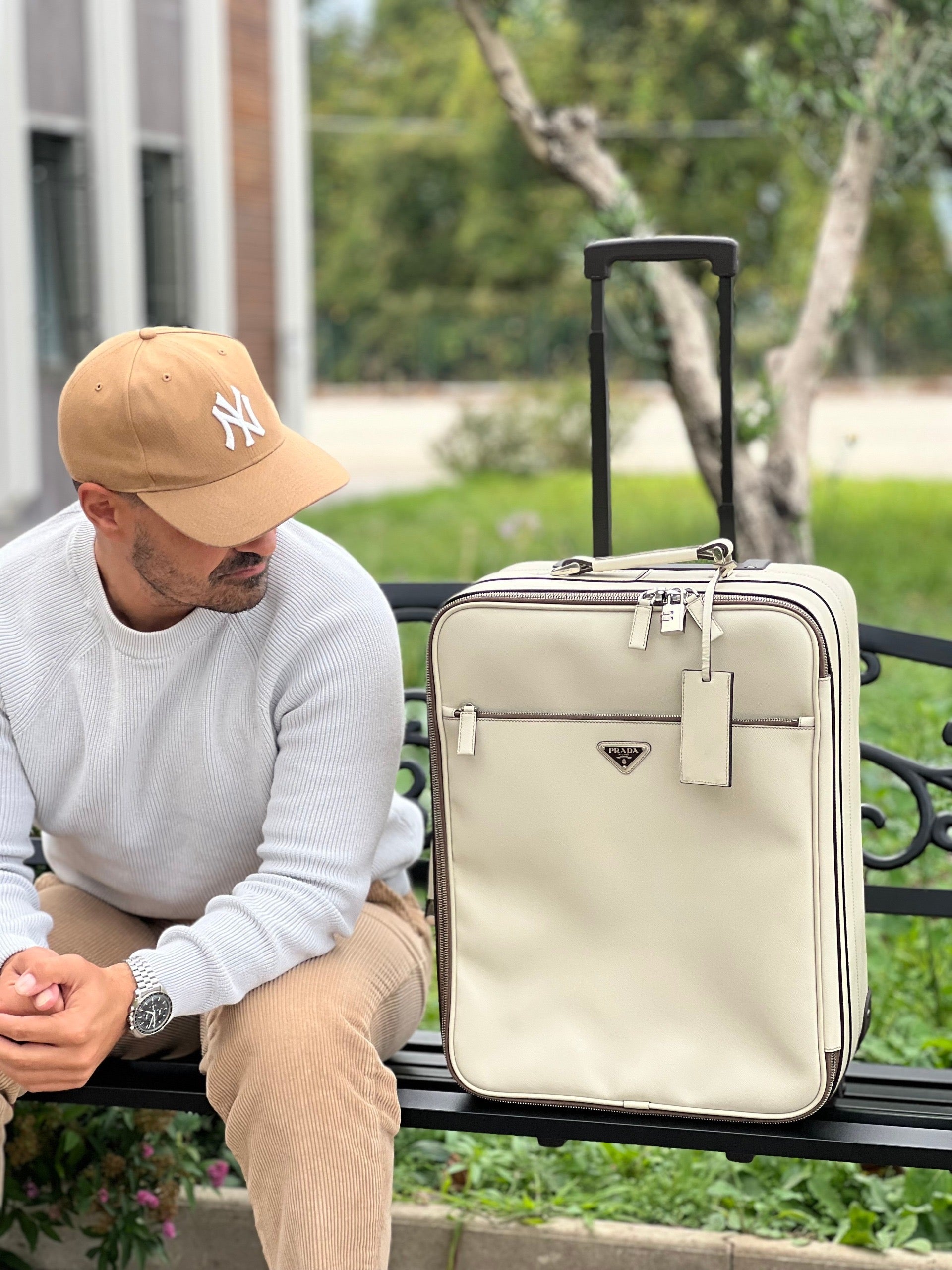 Ragazzo che mostra una valigia Prada in pelle saffiano bianco latte e finiture nere, con dettagli metallici argentati. L’interno è foderato in tessuto jacquard con motivo logo Prada, dotato di cinghie regolabili per fissare il contenuto e tasca con zip. Dispone di doppia chiusura con cerniera e lucchetto numerico in metallo argentato, manico superiore e laterale in pelle, maniglia estraibile e ruote posteriori per facilitarne il trasporto. Completa di dustbag, clochette porta address e card d'autenticità.