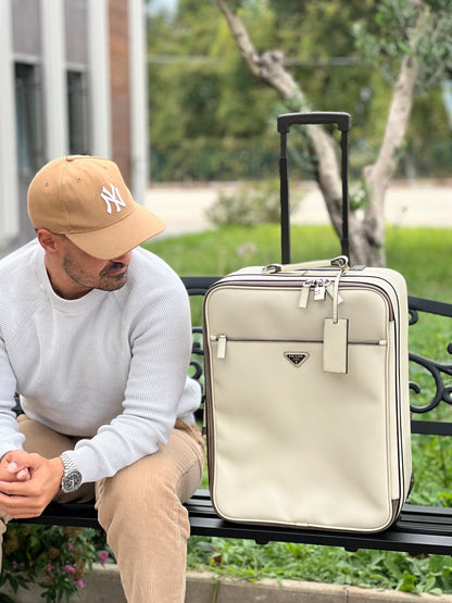 Ragazzo che mostra una valigia Prada in pelle saffiano bianco latte e finiture nere, con dettagli metallici argentati. L’interno è foderato in tessuto jacquard con motivo logo Prada, dotato di cinghie regolabili per fissare il contenuto e tasca con zip. Dispone di doppia chiusura con cerniera e lucchetto numerico in metallo argentato, manico superiore e laterale in pelle, maniglia estraibile e ruote posteriori per facilitarne il trasporto. Completa di dustbag, clochette porta address e card d'autenticità.