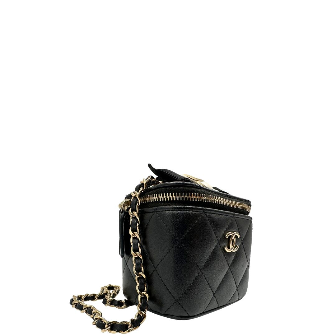 Laterale sinistro di una borsa Chanel in pelle matelassè nera rifinita con parti metalliche dorate; munita di una tracolla in pelle e catena intrecciata decorata con fiocco e camelia. Completa di scatola logata.