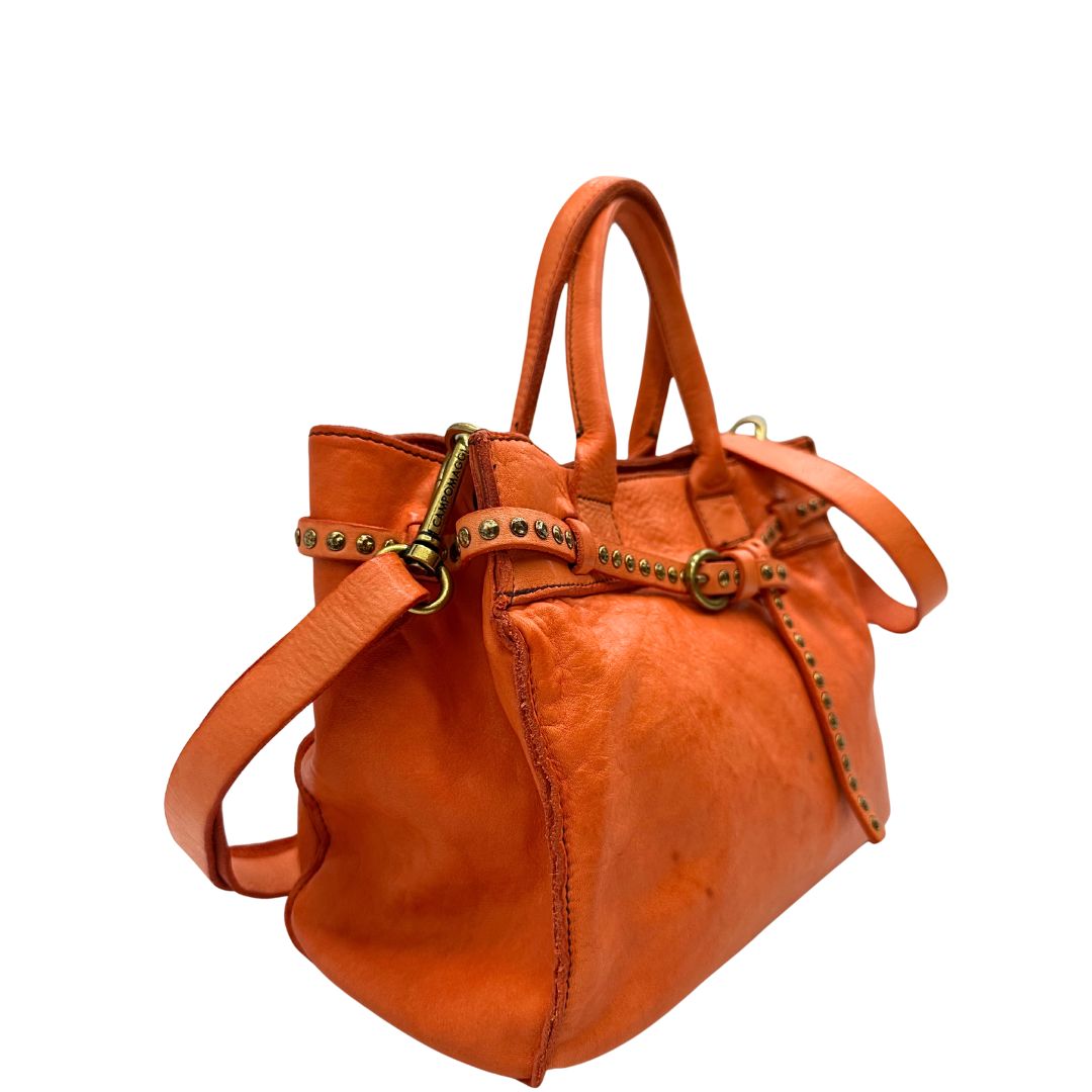 Laterale sinistro di una satchel Campomaggi in pelle arancione; munita di doppi manici e tracolla amovibile e regolabile. Parti metalliche dorate. Completa di dustbag e crema idratante per pelli.