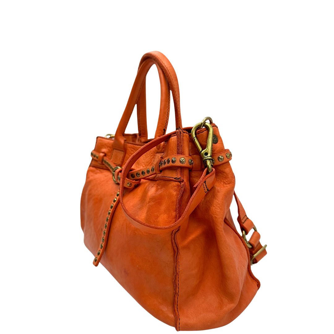 Laterale destro di una satchel Campomaggi in pelle arancione; munita di doppi manici e tracolla amovibile e regolabile. Parti metalliche dorate. Completa di dustbag e crema idratante per pelli.