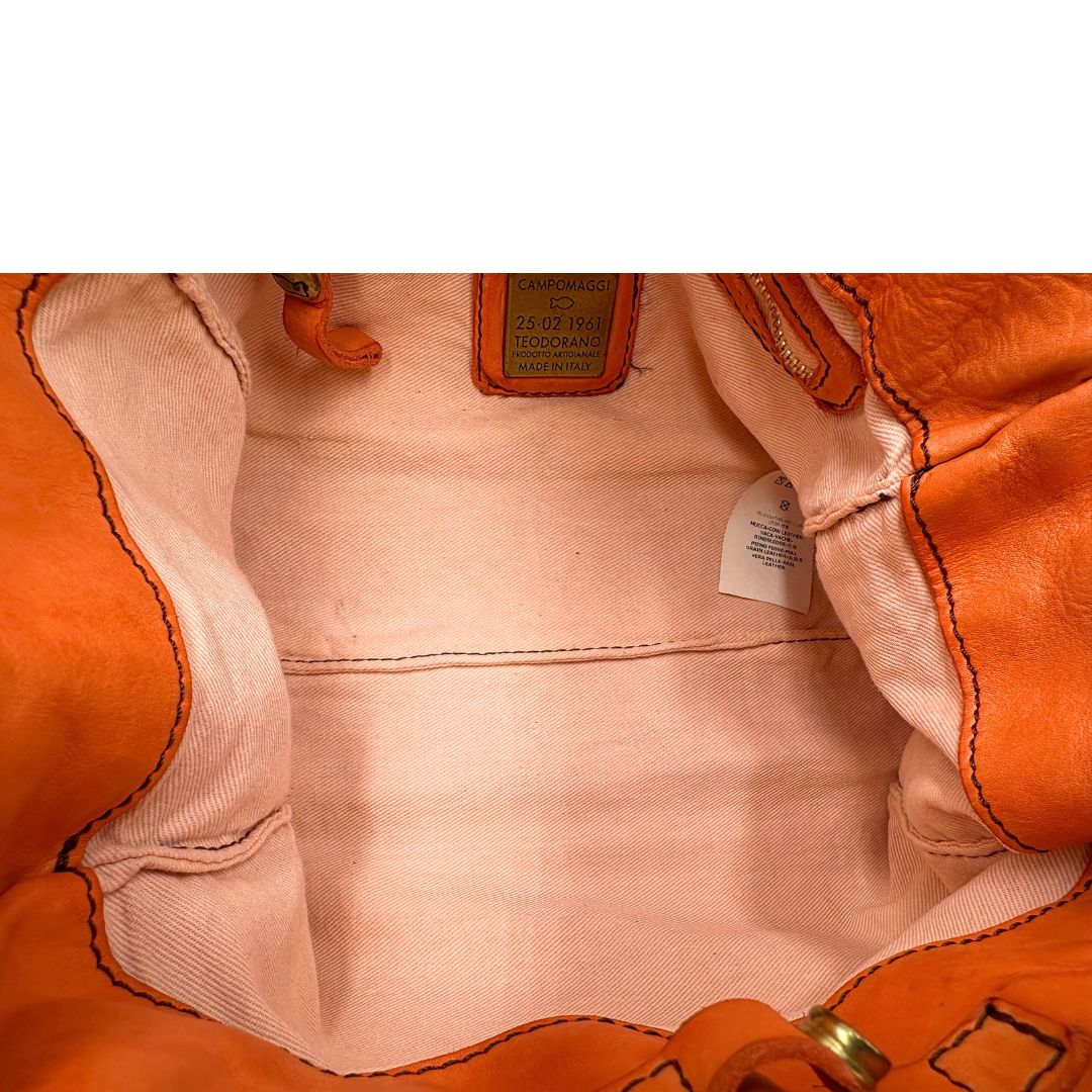 Interno di una satchel Campomaggi in pelle arancione; munita di doppi manici e tracolla amovibile e regolabile. Parti metalliche dorate. Completa di dustbag e crema idratante per pelli.