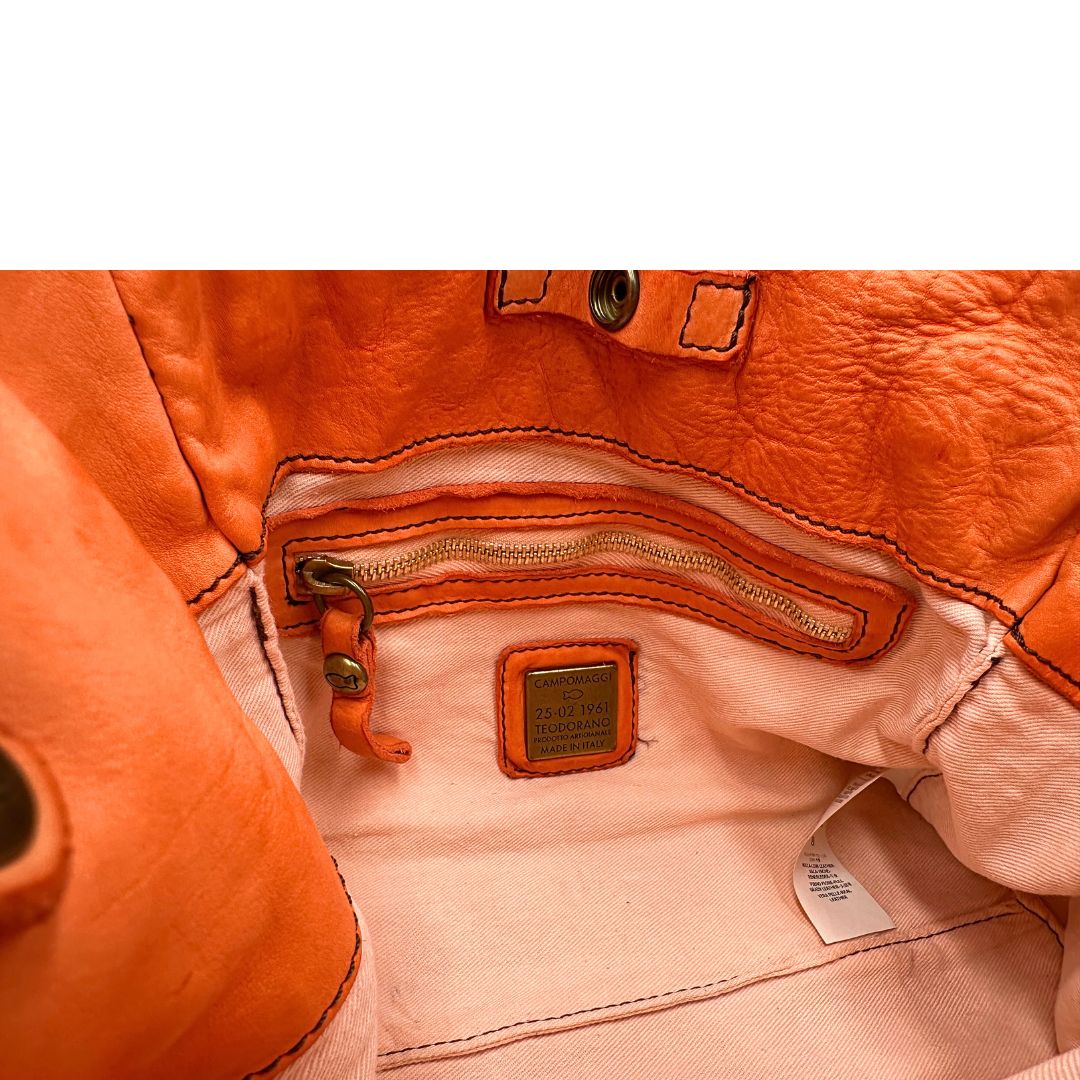 Interno di una satchel Campomaggi in pelle arancione; munita di doppi manici e tracolla amovibile e regolabile. Parti metalliche dorate. Completa di dustbag e crema idratante per pelli.