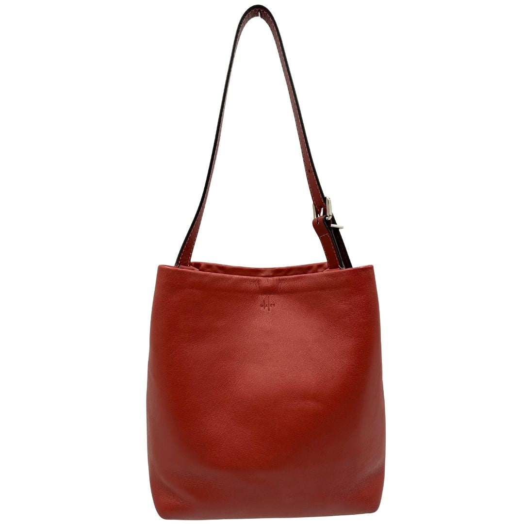 Front di una borsa a secchiello Hags in pelle color marrone bruciato con manico regolabile. Parti metalliche argentate. Indossabile a spalla o a tracolla. Completa di dustbag.