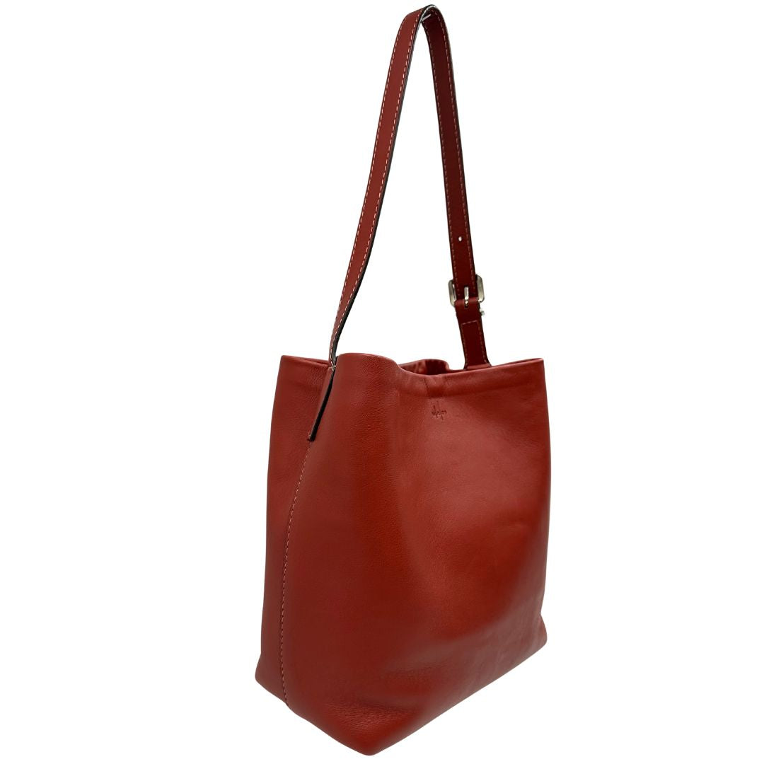 Laterale sinistro di una borsa a secchiello Hags in pelle color marrone bruciato con manico regolabile. Parti metalliche argentate. Indossabile a spalla o a tracolla. Completa di dustbag.