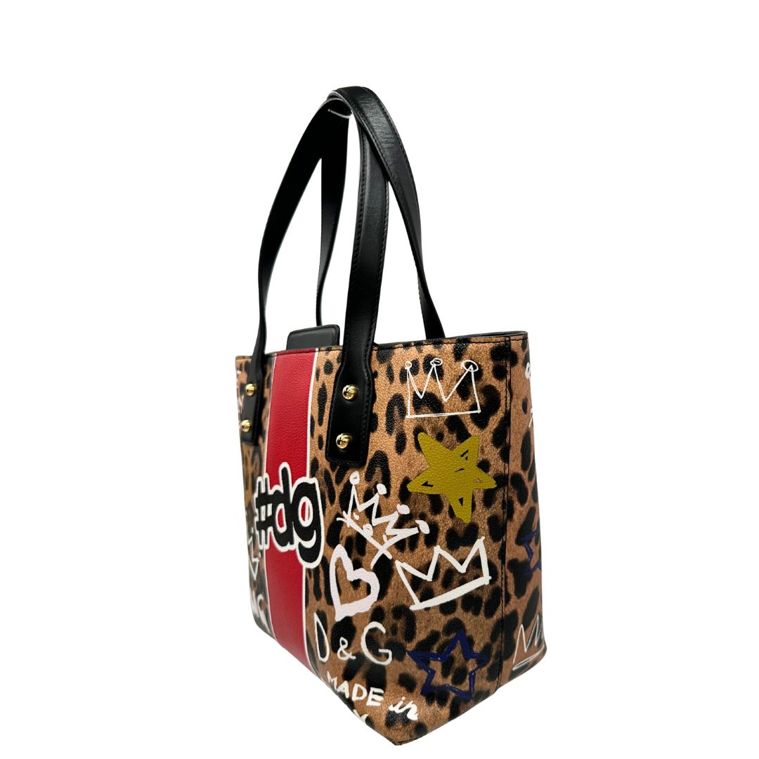 Laterale Borsa D&G tote Beatrice in canvas con motivo animalier e dettagli graffiti in stile street-art. Il design è arricchito da scritte, cuori e corone, e impreziosita da una striscia centrale rossa. Presenta doppi manici in pelle nera e parti metalliche dorate. Completa di dustbag, cartellino e card d'autenticità. Originale, usato, in condizioni ottime. 