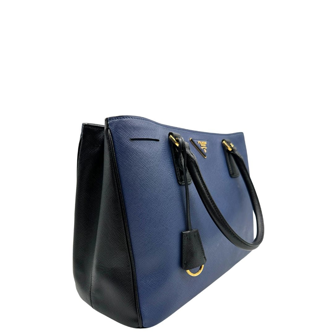 lato Borsa Prada in pelle saffiano blu e nera con parti metalliche dorate; munita di doppi manici stondati e impreziosita da un portachiavi. Completa di dustbag, di lusso, originale, ottime condizoni, usata. 