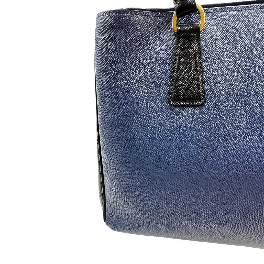 lato Borsa Prada in pelle saffiano blu e nera con parti metalliche dorate; munita di doppi manici stondati e impreziosita da un portachiavi. Completa di dustbag, di lusso, originale, ottime condizoni, usata. 