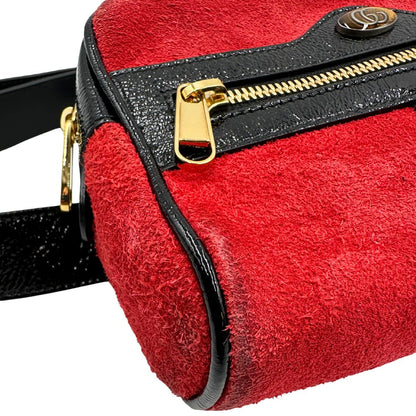 Angolo di un marsupio Gucci Ophidia in pelle scamosciata rossa con finiture in pelle verniciata nera, striscia Web centrale in tessuto blu e rosso, cintura regolabile e amovibile, parti metalliche dorate. Completo di dustbag.