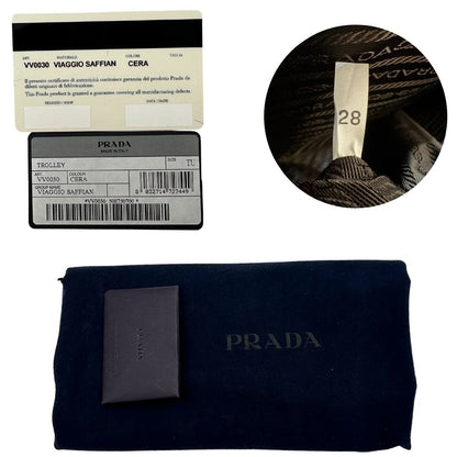 Dustbag e card di una valigia Prada in pelle saffiano bianco latte e finiture nere, con dettagli metallici argentati. L’interno è foderato in tessuto jacquard con motivo logo Prada, dotato di cinghie regolabili per fissare il contenuto e tasca con zip. Dispone di doppia chiusura con cerniera e lucchetto numerico in metallo argentato, manico superiore e laterale in pelle, maniglia estraibile e ruote posteriori per facilitarne il trasporto. Completa di dustbag, clochette porta address e card d'autenticità.