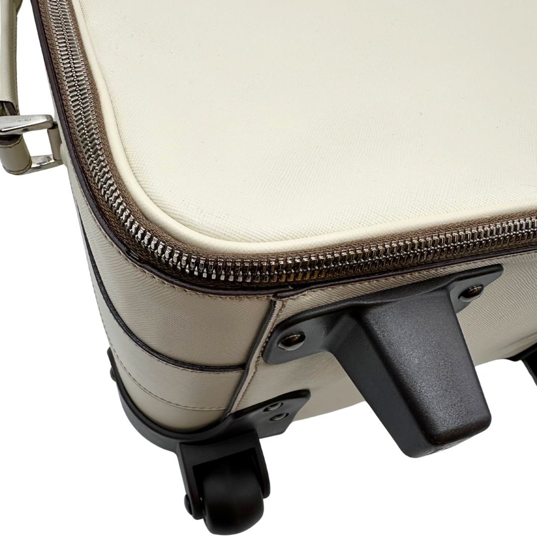 Appoggi di una valigia Prada in pelle saffiano bianco latte e finiture nere, con dettagli metallici argentati. L’interno è foderato in tessuto jacquard con motivo logo Prada, dotato di cinghie regolabili per fissare il contenuto e tasca con zip. Dispone di doppia chiusura con cerniera e lucchetto numerico in metallo argentato, manico superiore e laterale in pelle, maniglia estraibile e ruote posteriori per facilitarne il trasporto. Completa di dustbag, clochette porta address e card d'autenticità.