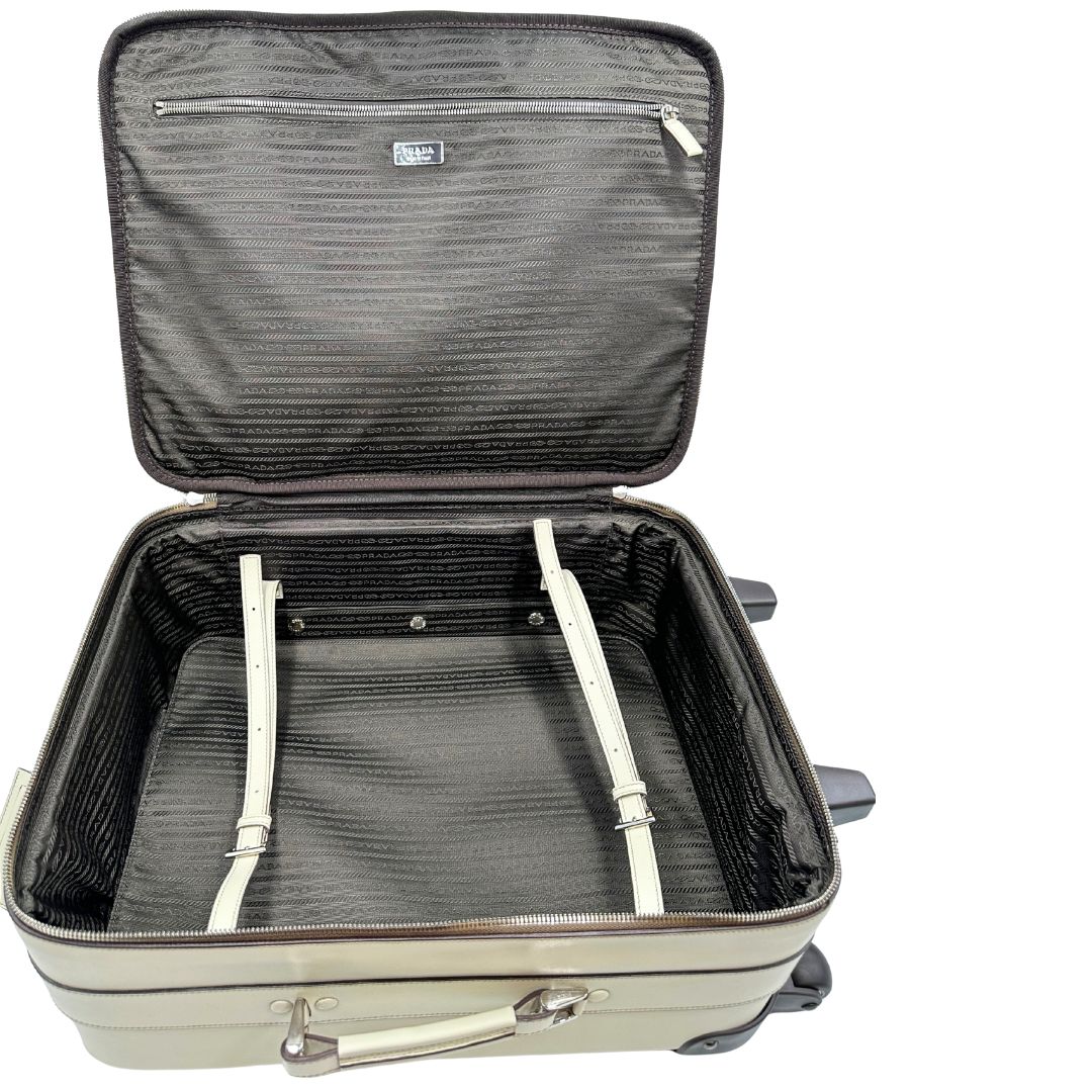 Interno di una valigia Prada in pelle saffiano bianco latte e finiture nere, con dettagli metallici argentati. L’interno è foderato in tessuto jacquard con motivo logo Prada, dotato di cinghie regolabili per fissare il contenuto e tasca con zip. Dispone di doppia chiusura con cerniera e lucchetto numerico in metallo argentato, manico superiore e laterale in pelle, maniglia estraibile e ruote posteriori per facilitarne il trasporto. Completa di dustbag, clochette porta address e card d'autenticità.