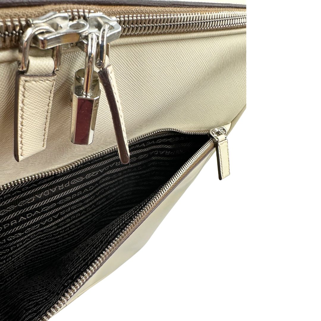Tasca cn zip di una valigia Prada in pelle saffiano bianco latte e finiture nere, con dettagli metallici argentati. L’interno è foderato in tessuto jacquard con motivo logo Prada, dotato di cinghie regolabili per fissare il contenuto e tasca con zip. Dispone di doppia chiusura con cerniera e lucchetto numerico in metallo argentato, manico superiore e laterale in pelle, maniglia estraibile e ruote posteriori per facilitarne il trasporto. Completa di dustbag, clochette porta address e card d'autenticità.
