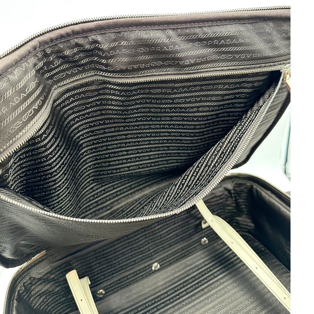 Interno di una valigia Prada in pelle saffiano bianco latte e finiture nere, con dettagli metallici argentati. L’interno è foderato in tessuto jacquard con motivo logo Prada, dotato di cinghie regolabili per fissare il contenuto e tasca con zip. Dispone di doppia chiusura con cerniera e lucchetto numerico in metallo argentato, manico superiore e laterale in pelle, maniglia estraibile e ruote posteriori per facilitarne il trasporto. Completa di dustbag, clochette porta address e card d'autenticità.