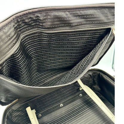Interno di una valigia Prada in pelle saffiano bianco latte e finiture nere, con dettagli metallici argentati. L’interno è foderato in tessuto jacquard con motivo logo Prada, dotato di cinghie regolabili per fissare il contenuto e tasca con zip. Dispone di doppia chiusura con cerniera e lucchetto numerico in metallo argentato, manico superiore e laterale in pelle, maniglia estraibile e ruote posteriori per facilitarne il trasporto. Completa di dustbag, clochette porta address e card d'autenticità.