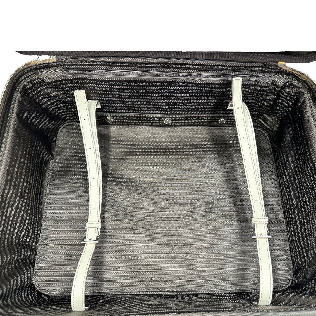 Interno di una valigia Prada in pelle saffiano bianco latte e finiture nere, con dettagli metallici argentati. L’interno è foderato in tessuto jacquard con motivo logo Prada, dotato di cinghie regolabili per fissare il contenuto e tasca con zip. Dispone di doppia chiusura con cerniera e lucchetto numerico in metallo argentato, manico superiore e laterale in pelle, maniglia estraibile e ruote posteriori per facilitarne il trasporto. Completa di dustbag, clochette porta address e card d'autenticità.