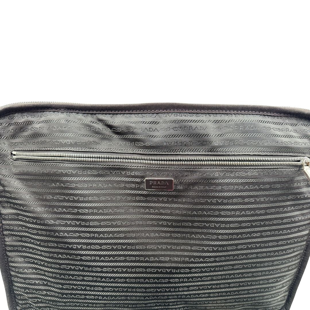 Interno di una valigia Prada in pelle saffiano bianco latte e finiture nere, con dettagli metallici argentati. L’interno è foderato in tessuto jacquard con motivo logo Prada, dotato di cinghie regolabili per fissare il contenuto e tasca con zip. Dispone di doppia chiusura con cerniera e lucchetto numerico in metallo argentato, manico superiore e laterale in pelle, maniglia estraibile e ruote posteriori per facilitarne il trasporto. Completa di dustbag, clochette porta address e card d'autenticità.