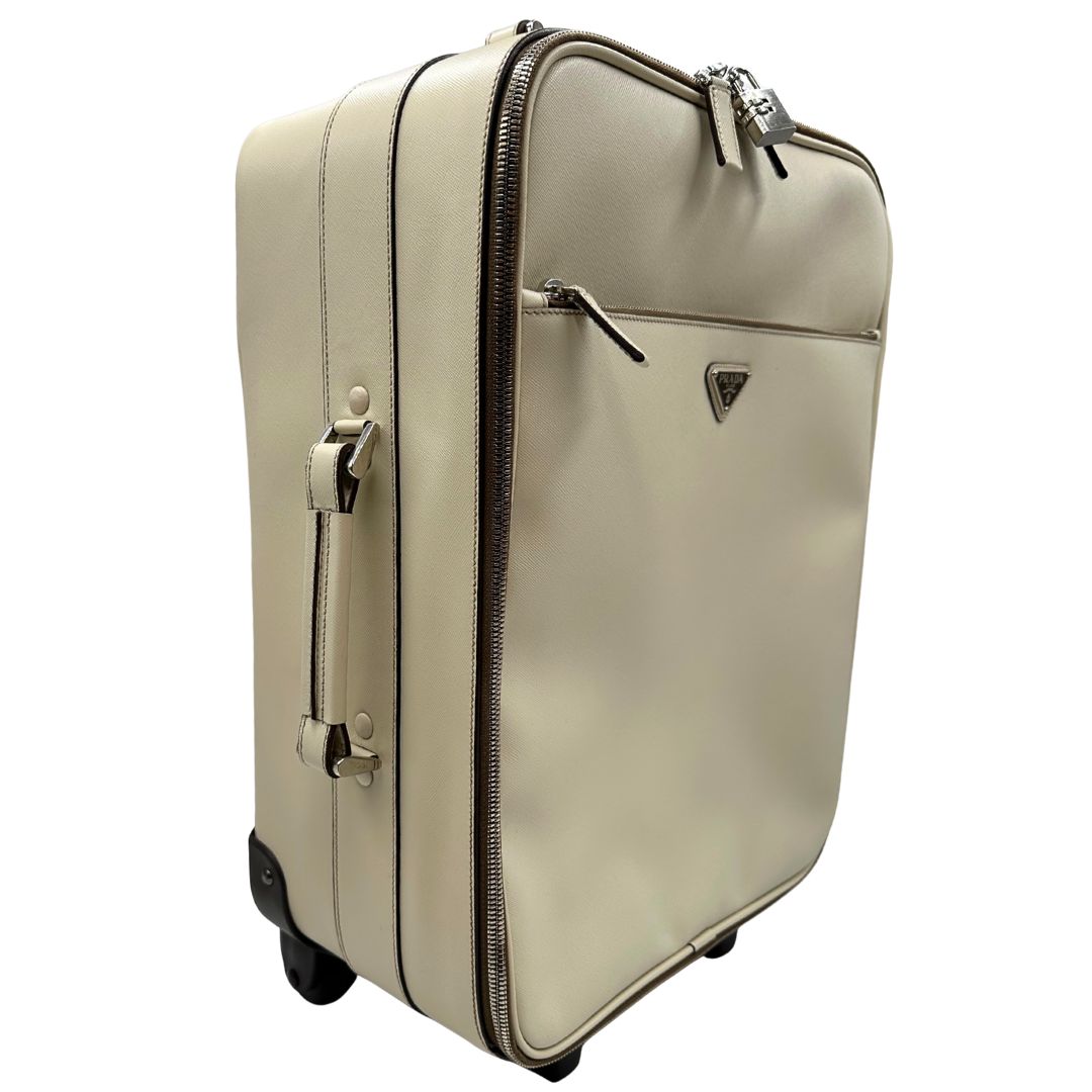 Laterale sinistro di una valigia Prada in pelle saffiano bianco latte e finiture nere, con dettagli metallici argentati. L’interno è foderato in tessuto jacquard con motivo logo Prada, dotato di cinghie regolabili per fissare il contenuto e tasca con zip. Dispone di doppia chiusura con cerniera e lucchetto numerico in metallo argentato, manico superiore e laterale in pelle, maniglia estraibile e ruote posteriori per facilitarne il trasporto. Completa di dustbag, clochette porta address e card d'autenticità.