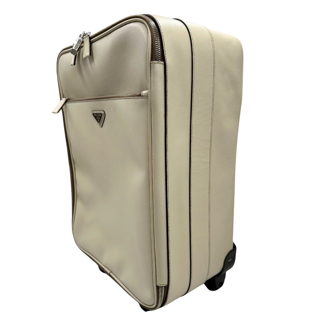 Laterale destro di una valigia Prada in pelle saffiano bianco latte e finiture nere, con dettagli metallici argentati. L’interno è foderato in tessuto jacquard con motivo logo Prada, dotato di cinghie regolabili per fissare il contenuto e tasca con zip. Dispone di doppia chiusura con cerniera e lucchetto numerico in metallo argentato, manico superiore e laterale in pelle, maniglia estraibile e ruote posteriori per facilitarne il trasporto. Completa di dustbag, clochette porta address e card d'autenticità.