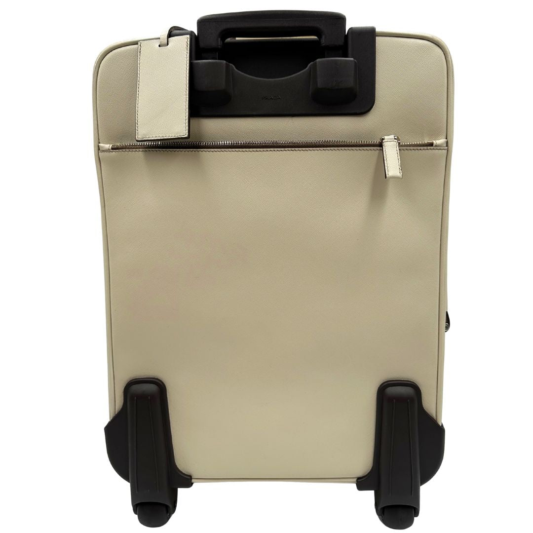 Retro di una valigia Prada in pelle saffiano bianco latte e finiture nere, con dettagli metallici argentati. L’interno è foderato in tessuto jacquard con motivo logo Prada, dotato di cinghie regolabili per fissare il contenuto e tasca con zip. Dispone di doppia chiusura con cerniera e lucchetto numerico in metallo argentato, manico superiore e laterale in pelle, maniglia estraibile e ruote posteriori per facilitarne il trasporto. Completa di dustbag, clochette porta address e card d'autenticità.