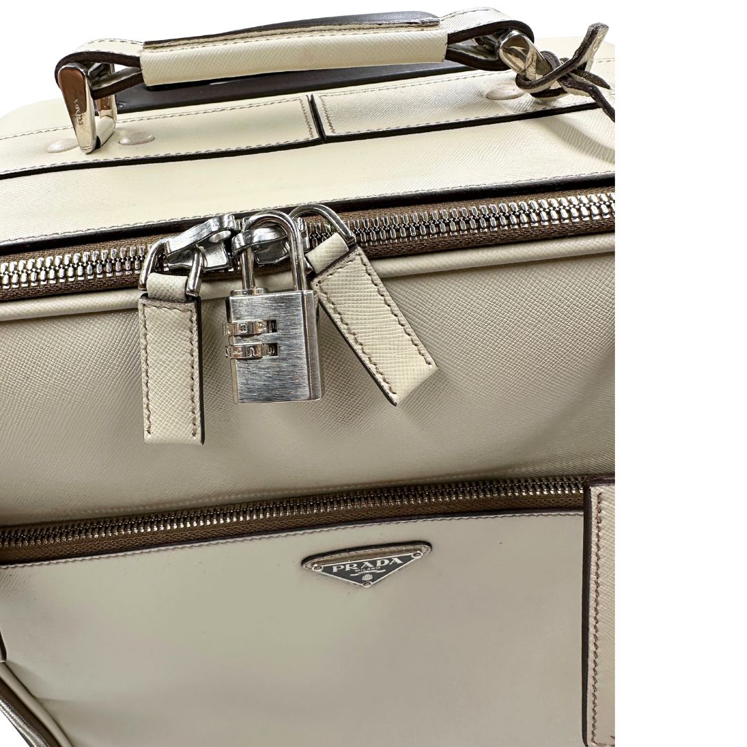 Chiusura di una valigia Prada in pelle saffiano bianco latte e finiture nere, con dettagli metallici argentati. L’interno è foderato in tessuto jacquard con motivo logo Prada, dotato di cinghie regolabili per fissare il contenuto e tasca con zip. Dispone di doppia chiusura con cerniera e lucchetto numerico in metallo argentato, manico superiore e laterale in pelle, maniglia estraibile e ruote posteriori per facilitarne il trasporto. Completa di dustbag, clochette porta address e card d'autenticità.