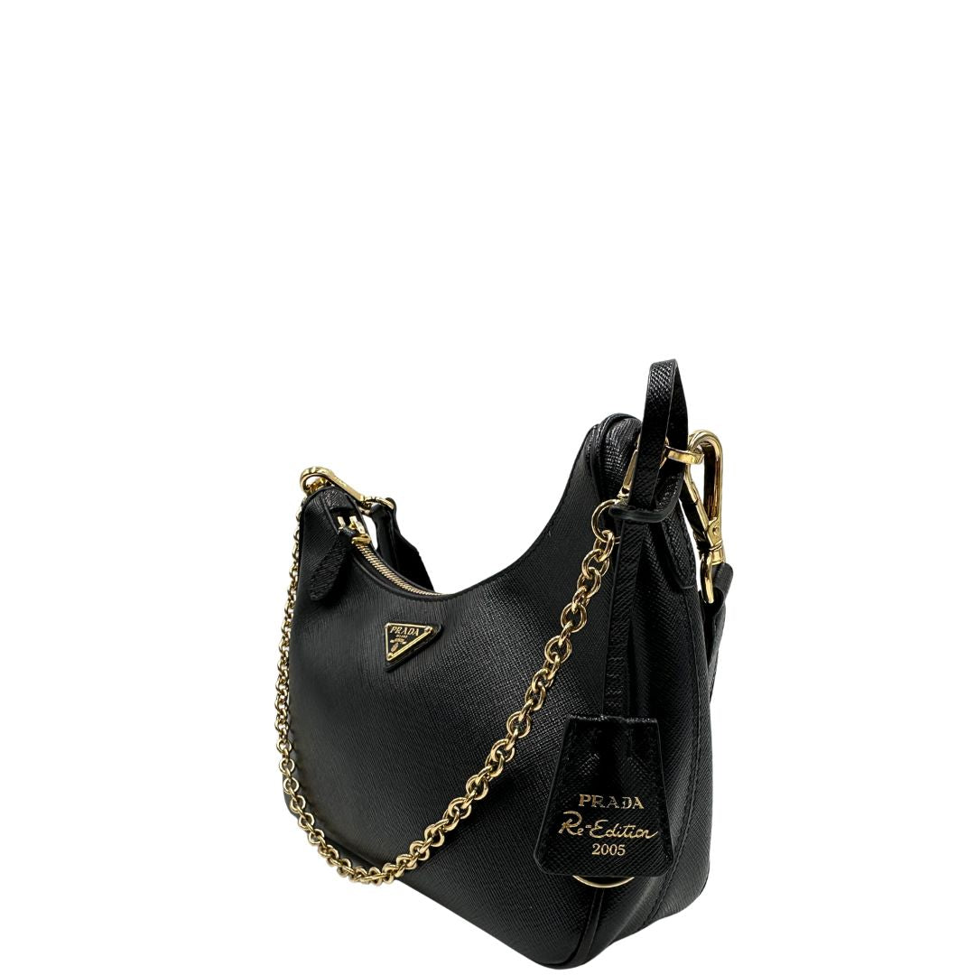 Laterale destro di una borsa Prada in pelle saffiano nera, con parti metalliche dorate. Dotata di una tracolla in tessuto regolabile ed amovibile, con pouch porta monete removibile e di una tracollina in catena. Munita di clochette porta chiavi. Indossabile a spalla o a tracolla. Full set.