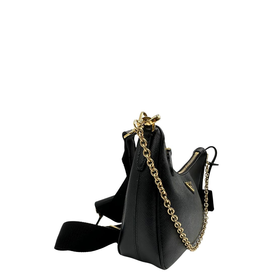 Laterale sinistro di una borsa Prada in pelle saffiano nera, con parti metalliche dorate. Dotata di una tracolla in tessuto regolabile ed amovibile, con pouch porta monete removibile e di una tracollina in catena. Munita di clochette porta chiavi. Indossabile a spalla o a tracolla. Full set.