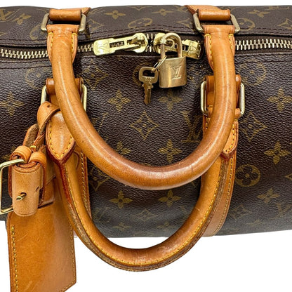 Manici di un Keepall Bandoulière in canvas monogram con finiture in vacchetta naturale e parti metalliche dorate; munito di doppi manici stondati e una tracolla amovibile e regolabile on spallaccio. Completo di lucchetto con chiave, tag porta address e ferma manici.