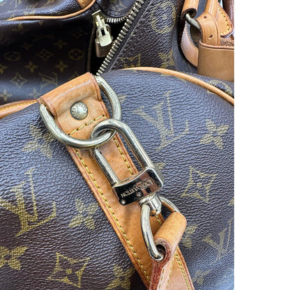 Gancio della tracolla di un Keepall Bandoulière in canvas monogram con finiture in vacchetta naturale e parti metalliche dorate; munito di doppi manici stondati e una tracolla amovibile e regolabile on spallaccio. Completo di lucchetto con chiave, tag porta address e ferma manici.