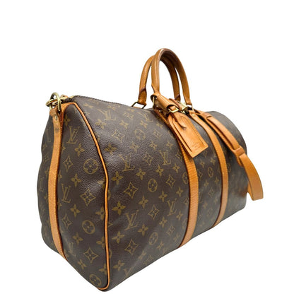 Laterale sinistro di un Keepall Bandoulière in canvas monogram con finiture in vacchetta naturale e parti metalliche dorate; munito di doppi manici stondati e una tracolla amovibile e regolabile on spallaccio. Completo di lucchetto con chiave, tag porta address e ferma manici.