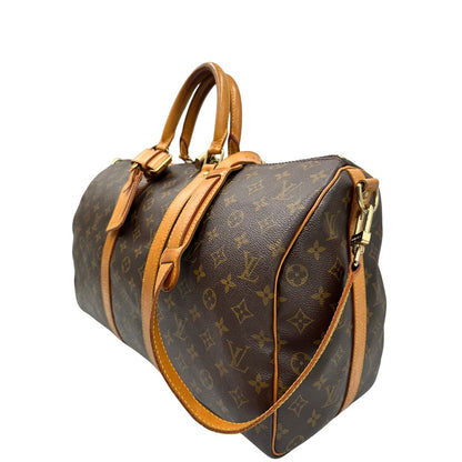 Laterale destro di un Keepall Bandoulière in canvas monogram con finiture in vacchetta naturale e parti metalliche dorate; munito di doppi manici stondati e una tracolla amovibile e regolabile on spallaccio. Completo di lucchetto con chiave, tag porta address e ferma manici.