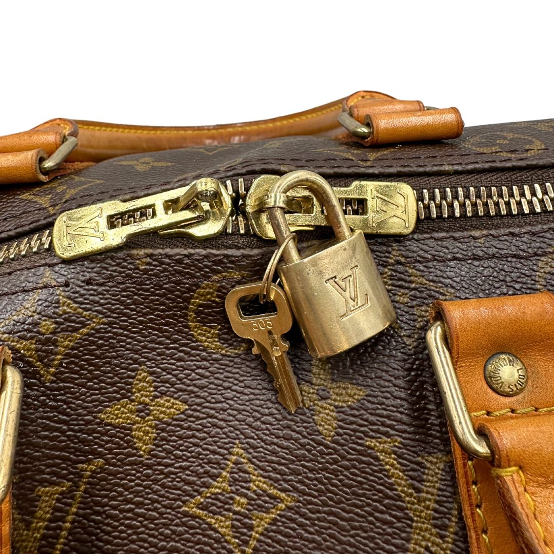 Dettagli metallici di un Keepall Bandoulière in canvas monogram con finiture in vacchetta naturale e parti metalliche dorate; munito di doppi manici stondati e una tracolla amovibile e regolabile on spallaccio. Completo di lucchetto con chiave, tag porta address e ferma manici.