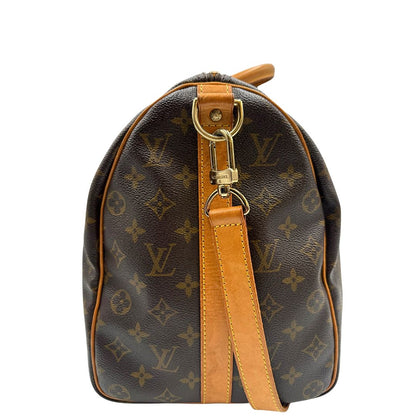 Vista laterale di un Keepall Bandoulière in canvas monogram con finiture in vacchetta naturale e parti metalliche dorate; munito di doppi manici stondati e una tracolla amovibile e regolabile on spallaccio. Completo di lucchetto con chiave, tag porta address e ferma manici.