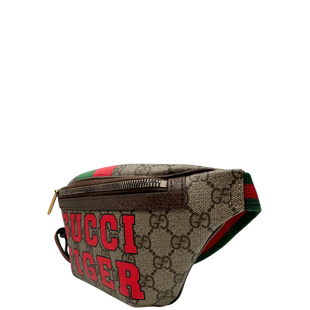 Laterale destro di un marsupio Gucci in canvas beige GG con metalleria dorata e finiture in pelle marroni. Stampa Gucci Tiger e fascia Web su front e retro. Tracolla in tessuto Web verde/rosso, regolabile. Completo di dustbag. Indossabile a marsupio o crossbody.