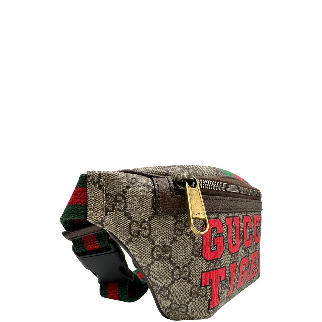 Laterale sinistro di un marsupio Gucci in canvas beige GG con metalleria dorata e finiture in pelle marroni. Stampa Gucci Tiger e fascia Web su front e retro. Tracolla in tessuto Web verde/rosso, regolabile. Completo di dustbag. Indossabile a marsupio o crossbody.
