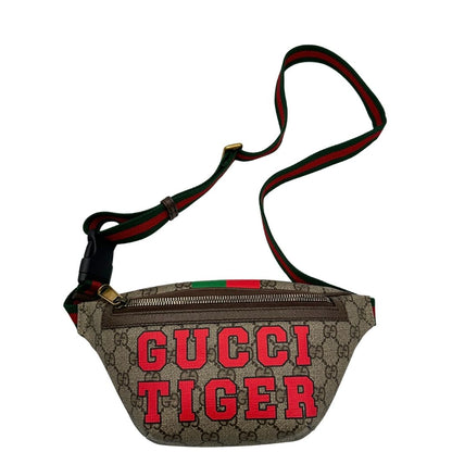 Front di un marsupio Gucci in canvas beige GG con metalleria dorata e finiture in pelle marroni. Stampa Gucci Tiger e fascia Web su front e retro. Tracolla in tessuto Web verde/rosso, regolabile. Completo di dustbag. Indossabile a marsupio o crossbody.