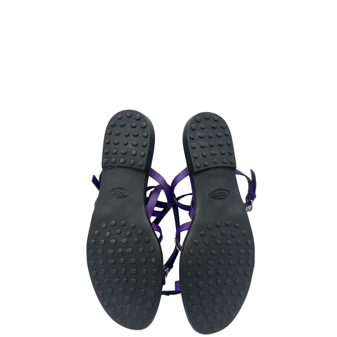 Suola di sandali Tod’s in pelle viola con fasce incrociate e borchie metalliche argentate. Presentano una chiusura con cinturino e la suola con i classici gommini in gomma. Completi di scatola e dustbag.