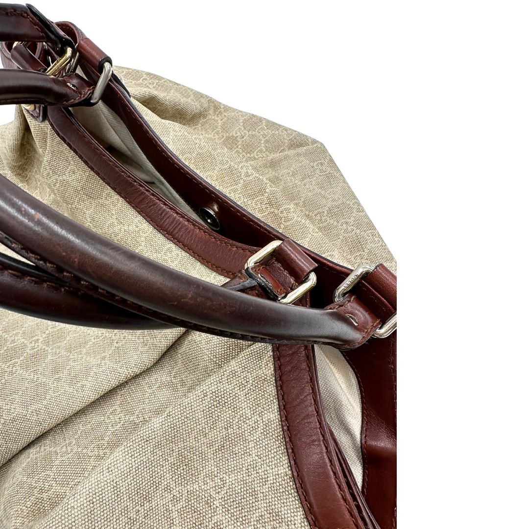 Manici di una borsa Gucci Sukey in tessuto beige GG con finiture in pelle marrone mogano, doppi manici stondati e ciondolo con doppie G incrociate. Rifinita con parti metalliche dorate. Indossabile a spalla.